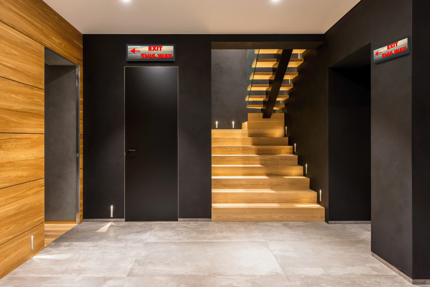 A lobby with dark walls and brown stairs with a POE panoramic stretch display that says "exit this way" above the door and on the wall next to a hallway.